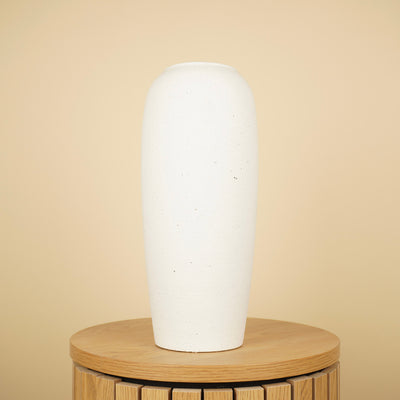 Hoge, slanke crèmekleurige vaas met matte afwerking en fijne spikkels op een ronde houten tafel.