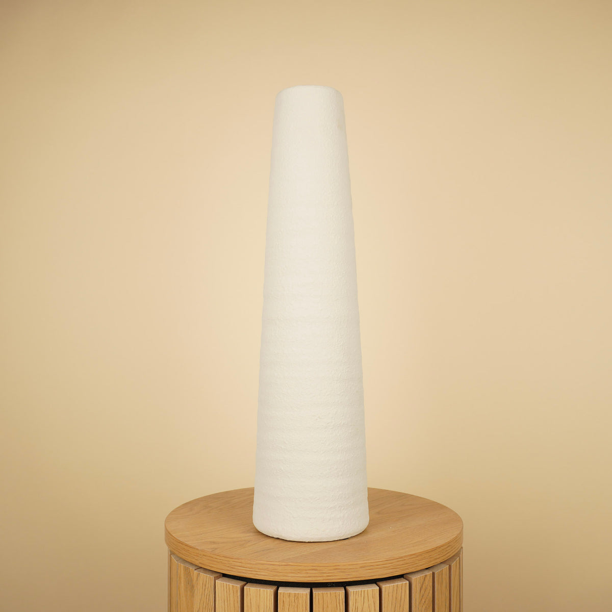 Hoge, slanke witte vaas met taps toelopende vorm en ruwe structuur op een ronde houten tafel.